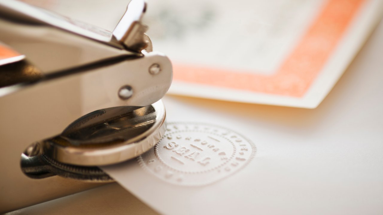 A metal seal press is stamping a circular embossed seal onto a piece of paper.