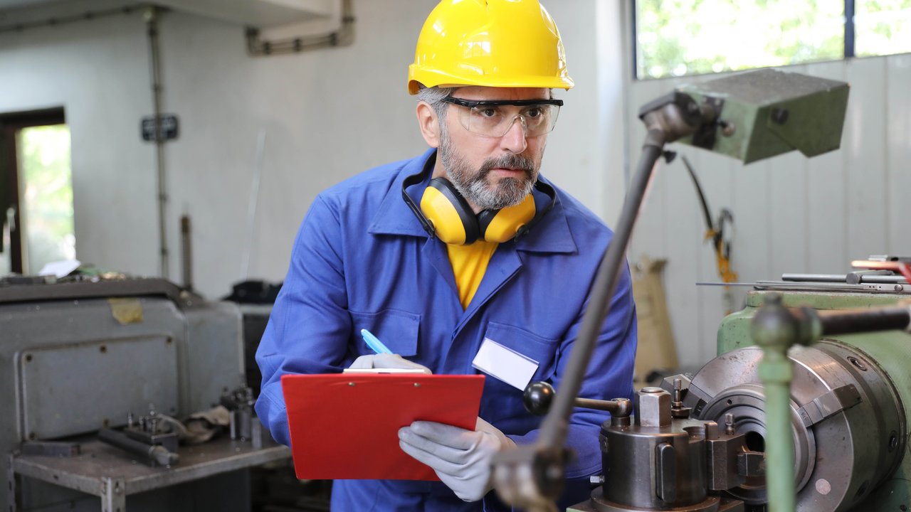 A man in a blue work uniform and yellow hard hat inspects machinery while taking notes on a clipboard.