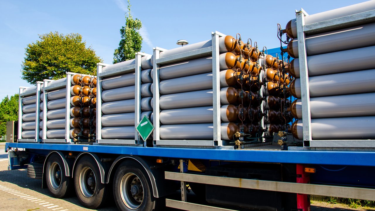 A large blue truck is parked on a paved area, carrying multiple rows of cylindrical gas tanks. The tanks are arranged in two distinct sections: one with silver cylinders and another with brown ones, both secured in metal frames. A green hazard sign is visible on the side, indicating the cargo's nature. Lush trees and a clear blue sky provide a vibrant backdrop.