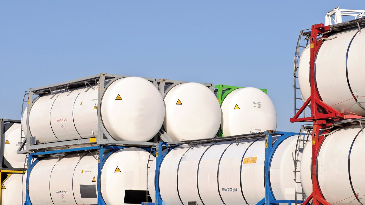 A stack of large, cylindrical storage tanks is displayed against a clear blue sky. The tanks are primarily white, with some featuring colored frames in blue, red, and green. Warning symbols are visible, indicating caution. Labels such as "FOODSTUFF ONLY" suggest specific contents, emphasizing safety and regulation.