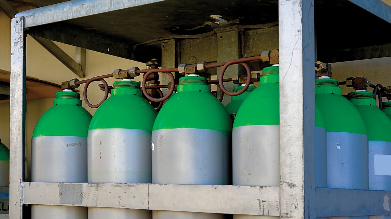 A row of metallic gas cylinders is secured in a sturdy metal rack. Each cylinder features a silver body with a bright green top, indicating their contents. Connecting pipes and valves are visible, suggesting a system for gas distribution. The background is neutral, emphasizing the industrial setting.