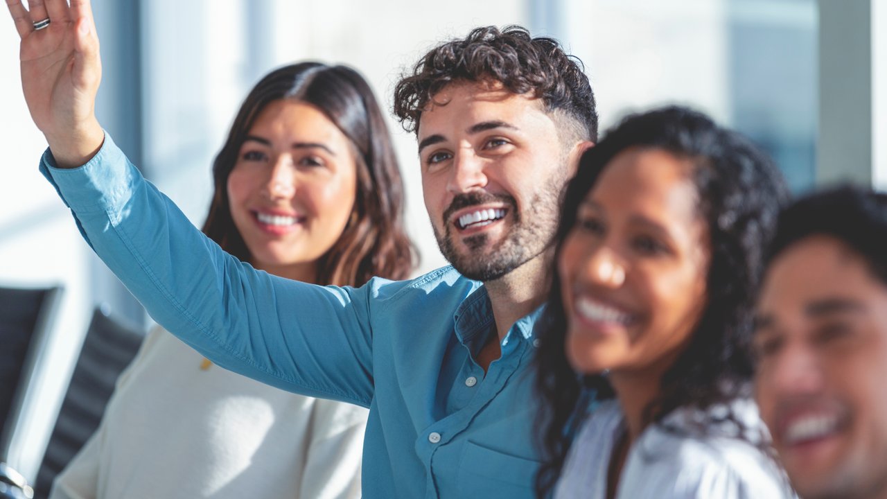 Group of people listening to a presentation. One person has their hand raised. They are at a seminar or meeting sitting at a table. They are all looking to the front of the room. Men and women dressed in casual business attire. Multi-cultural group with African, Latino and Caucasian.