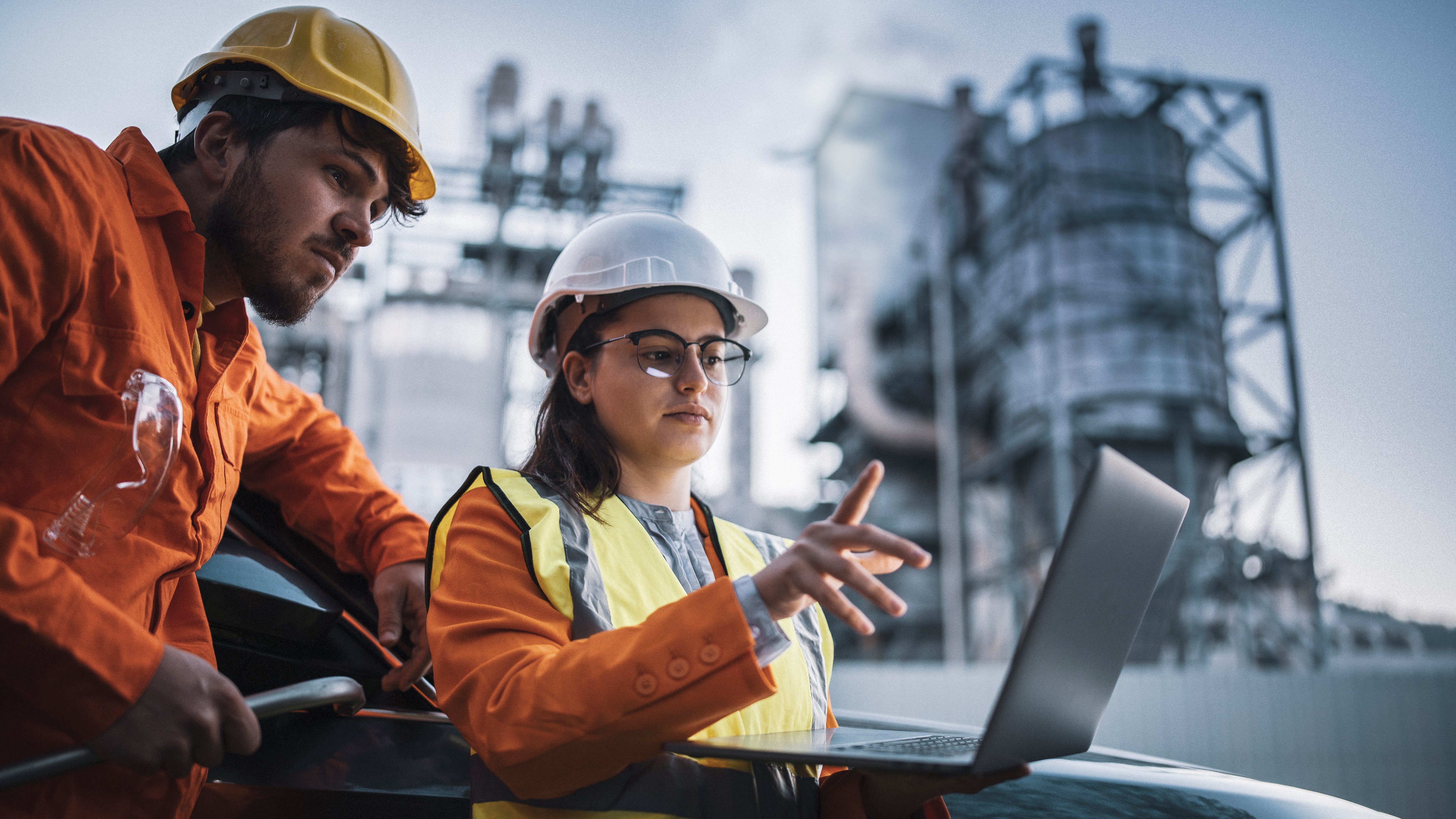 Two workers in safety gear discuss over a laptop near industrial buildings.
