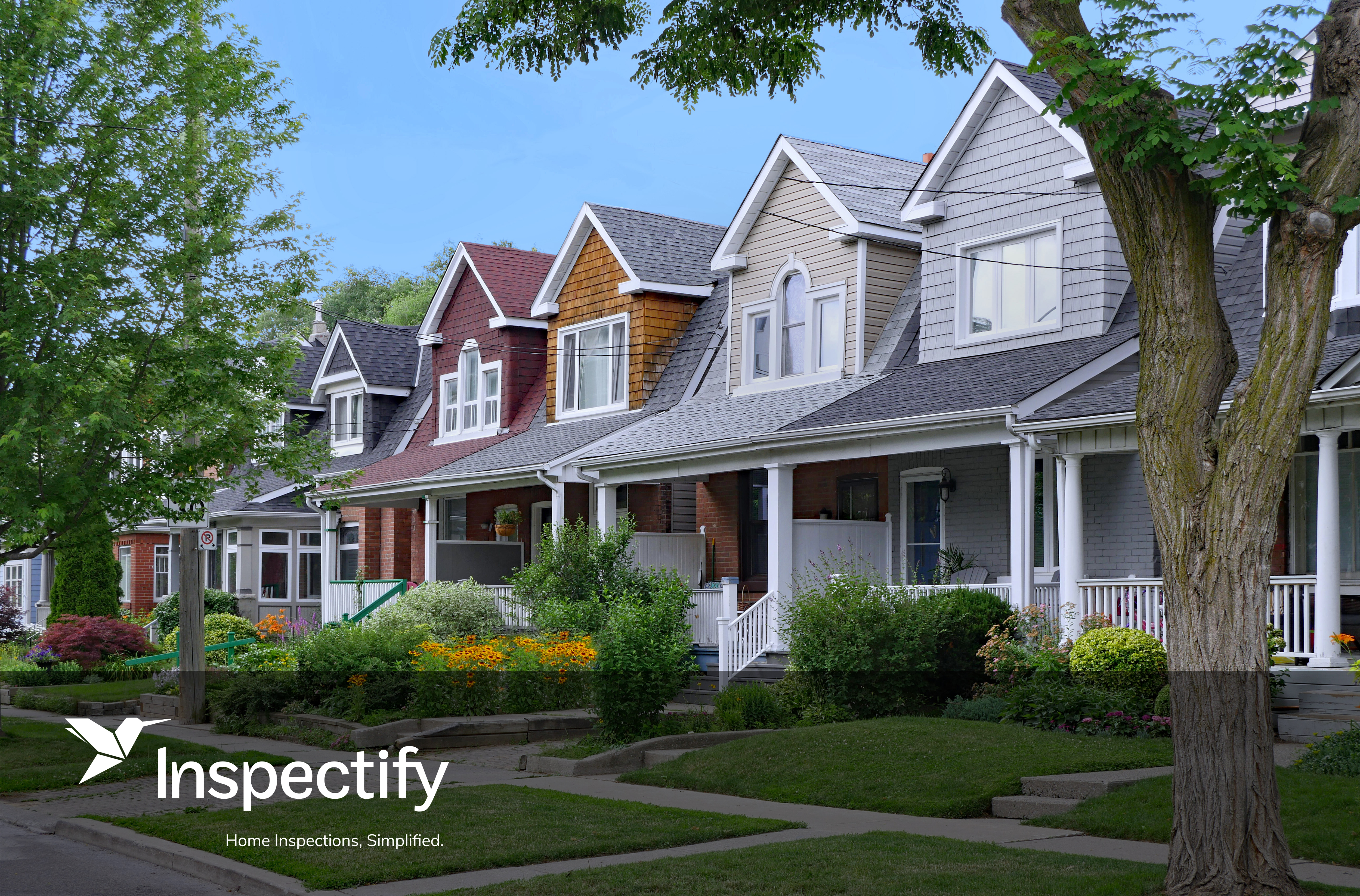 A row of charming houses features varied architectural styles, showcasing gabled roofs and inviting porches. Lush greenery and colorful flower beds enhance the front yards, creating a vibrant atmosphere. A clear blue sky provides a bright backdrop, while a utility pole subtly marks the neighborhood's edge.