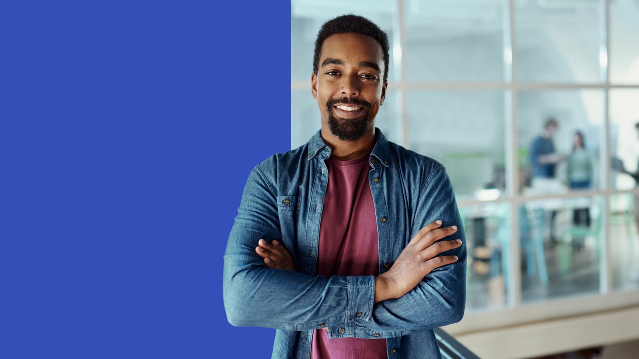 A person stands confidently with arms crossed, wearing a denim jacket over a maroon shirt. The background features a bright blue section alongside a modern workspace with blurred figures engaged in conversation. The overall atmosphere conveys professionalism and collaboration.