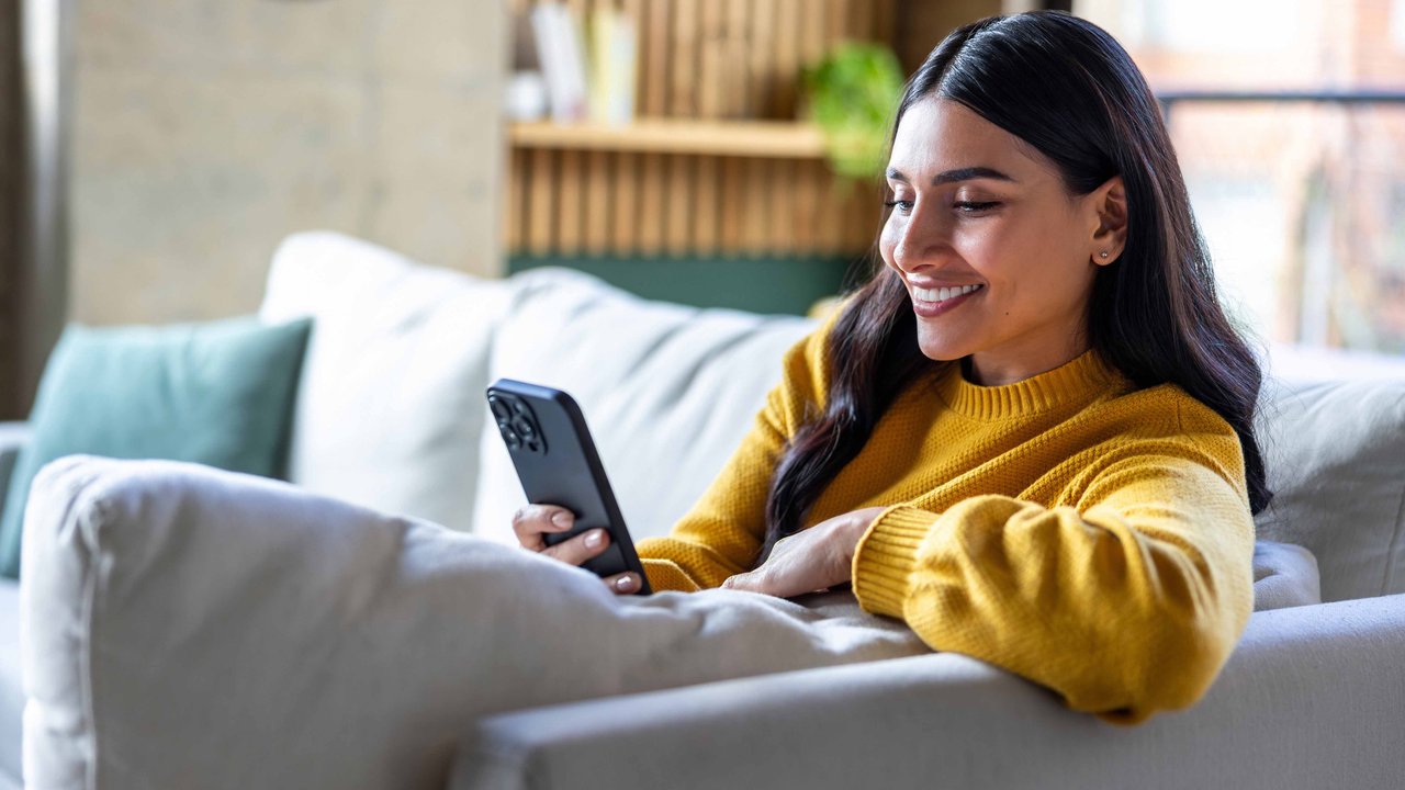 A person in a yellow sweater sits on a couch, holding a smartphone, with a cozy interior in the background.