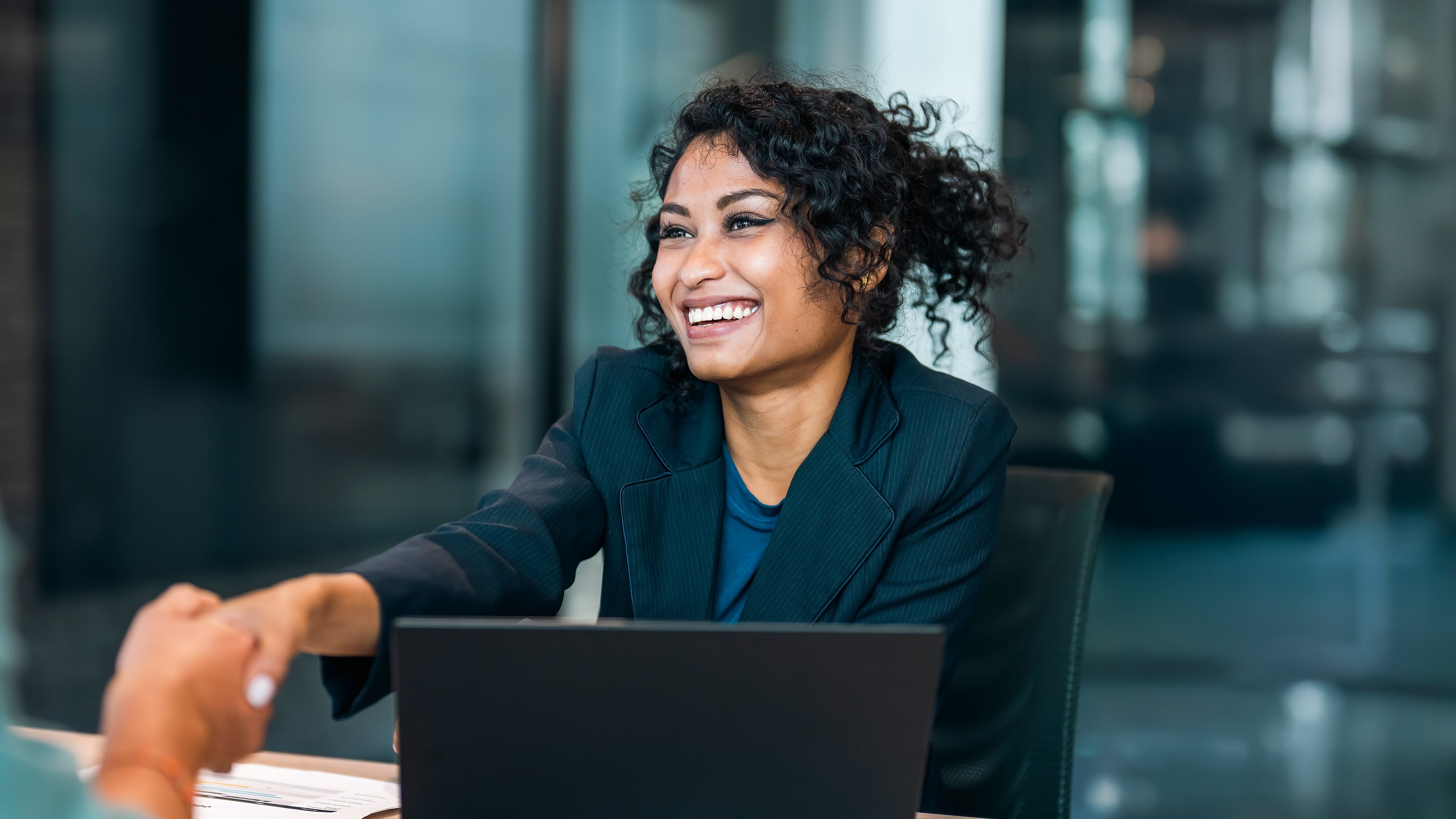 HSB target markets A person in a dark blazer sits at a conference table, reaching out to shake hands with someone off-camera. A laptop is open in front of them, and the background features large windows, suggesting a modern office environment. The atmosphere appears professional and collaborative.