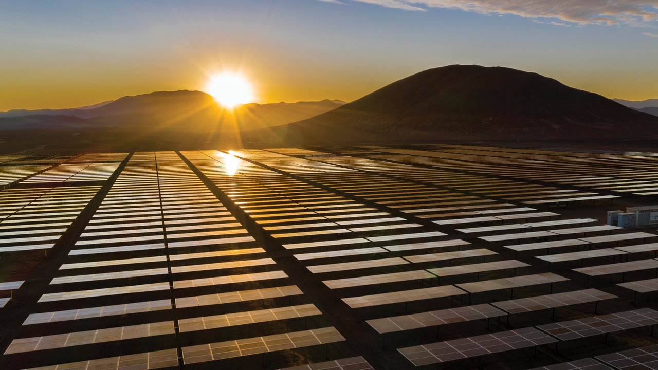 A solar farm with rows of solar panels under a sunrise and a mountain in the background.