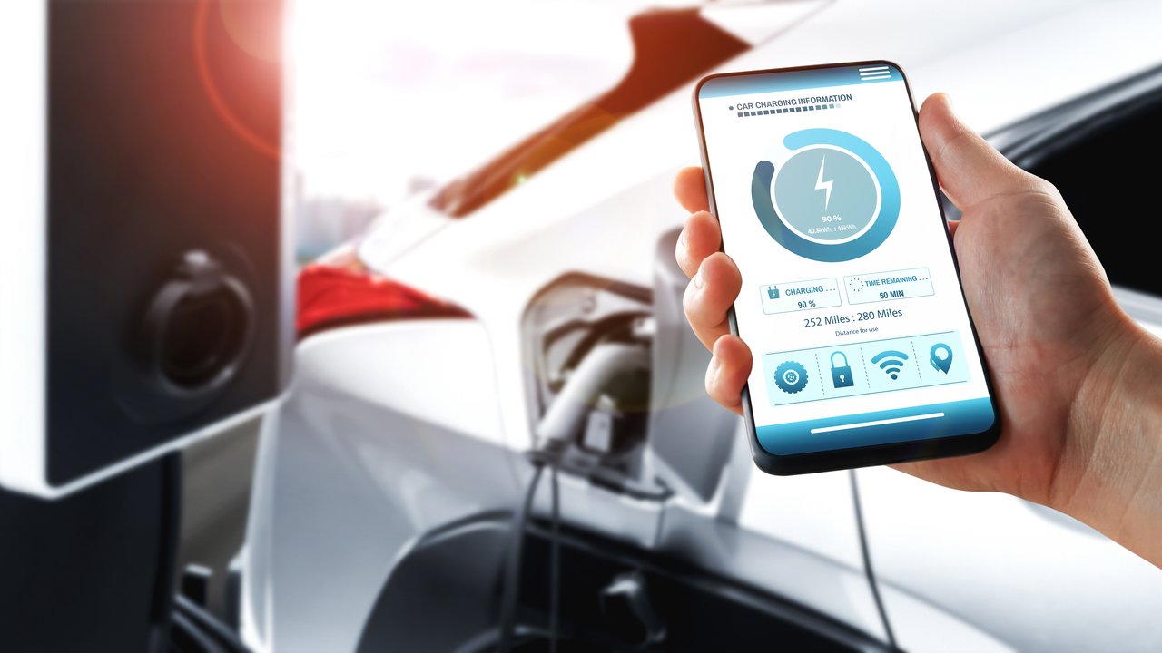 A hand holds a smartphone displaying electric vehicle charging information next to a charging station.