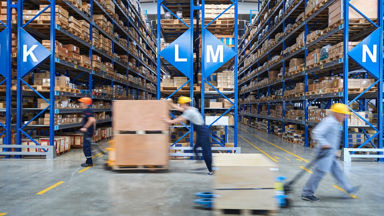 Warehouse interior with shelves of boxes; workers moving pallets with hand trucks.