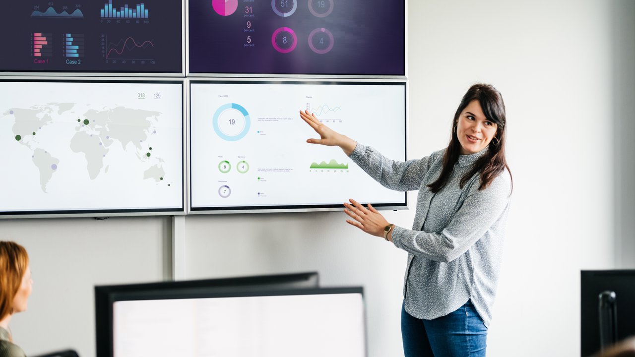 A businesswoman explaining series of graphs and data sets displayed on some large, wall mounted monitors in the office.