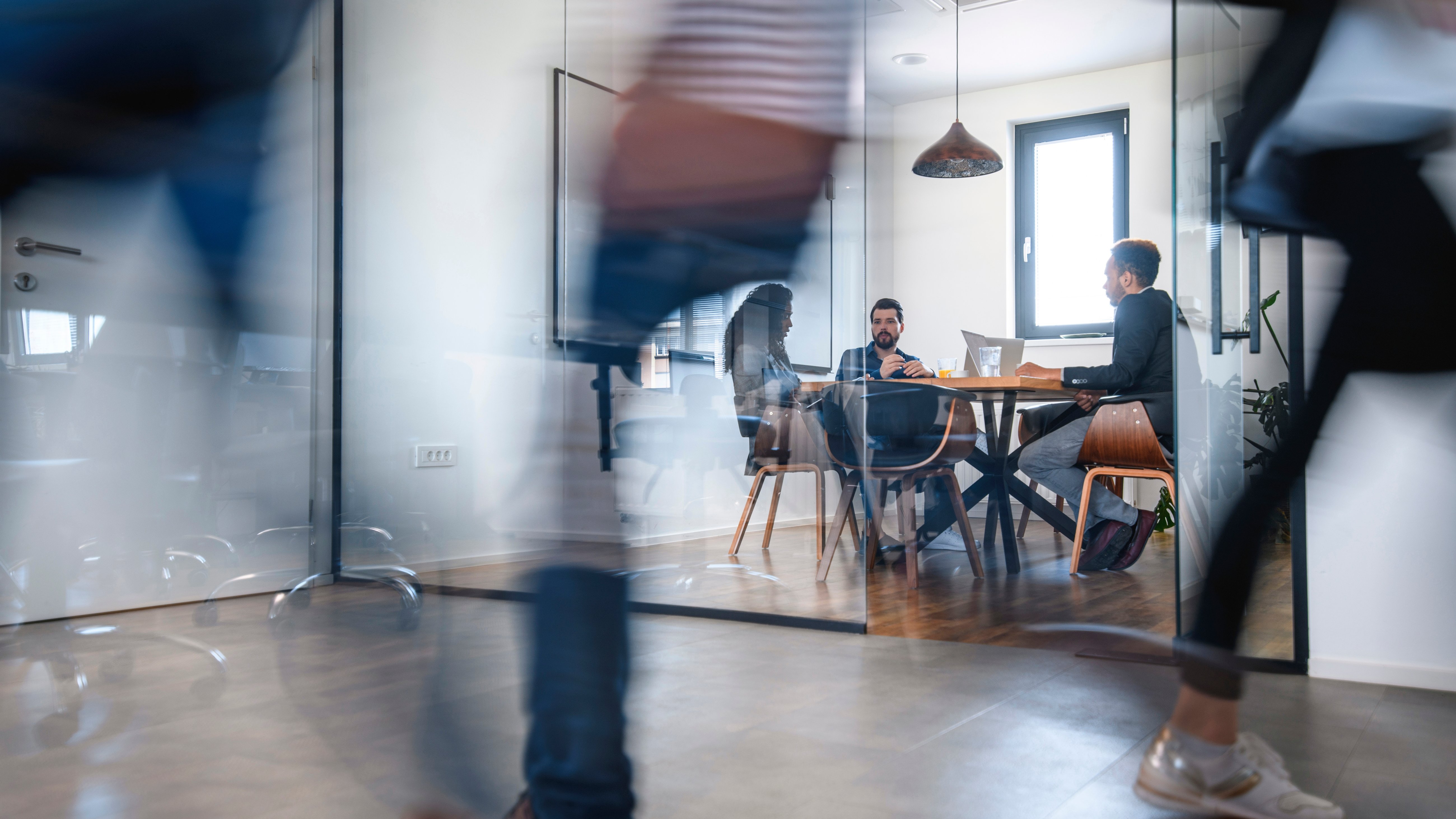 Blurred motion of colleagues walking briskly down office hallway as colleagues sit in conference room discussing ideas.