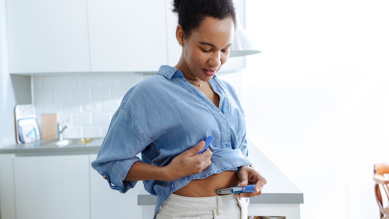 Woman administering GLP-1 shot in a kitchen A person stands in a bright kitchen, wearing a light blue button-up shirt and light-colored pants. They are lifting their shirt slightly, holding a blue GLP-1 device near their abdomen. The kitchen features modern appliances and a clean, minimalist design, with a glimpse of colorful items in a nearby cabinet.