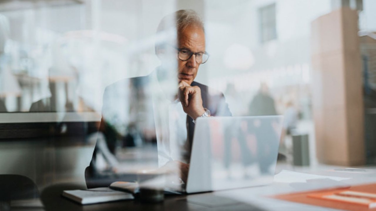 A person in a suit sits at a table, working on a laptop in a bright, modern office environment.