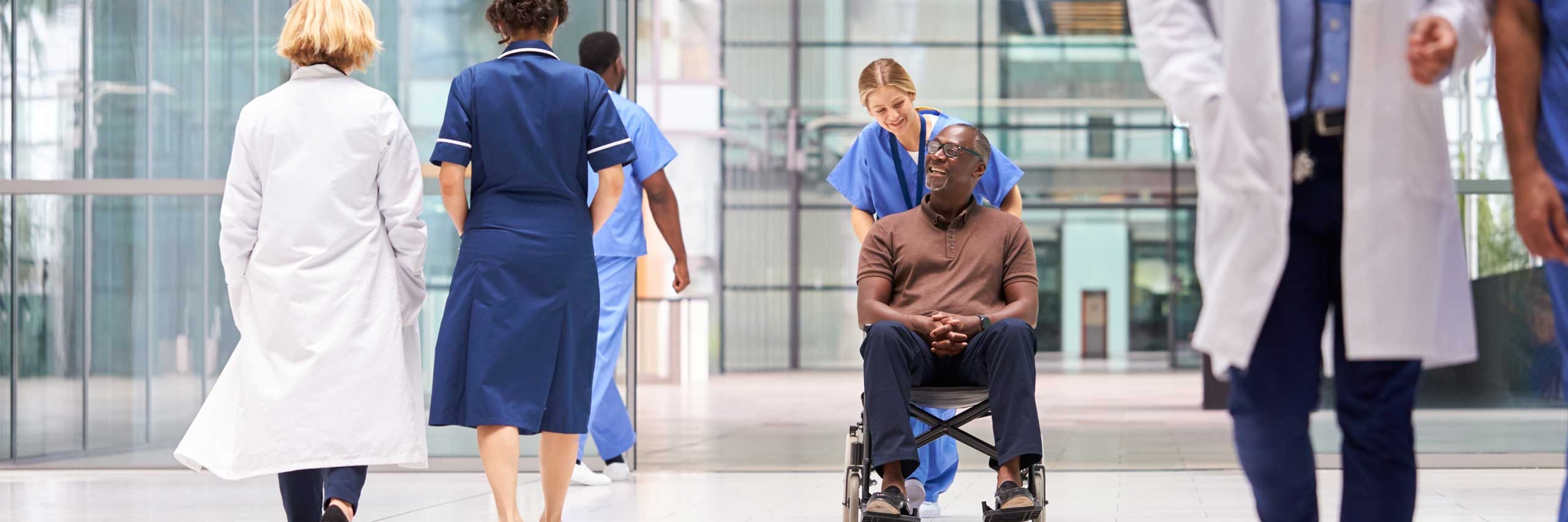Female nurse wearing scrubs wheeling patient in wheelchair through lobby of modern hospital building
