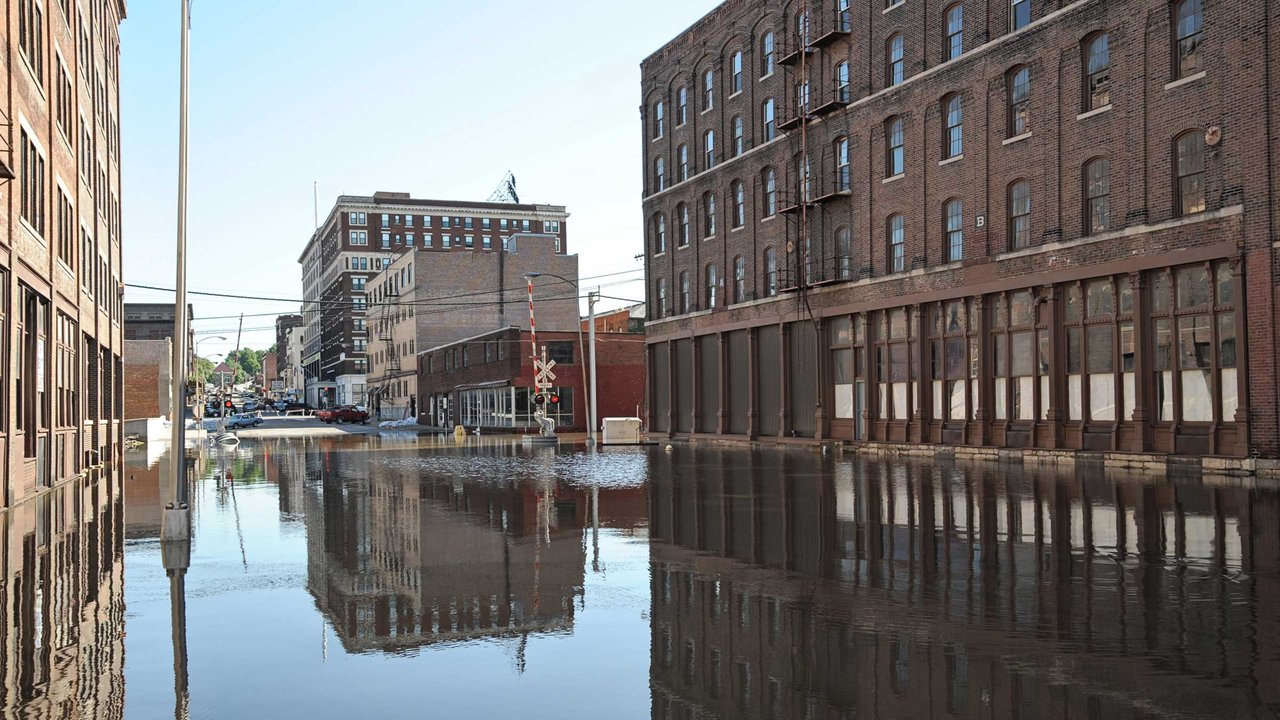 Flooded street with brick buildings and reflections in the water, clear sky in the background.
