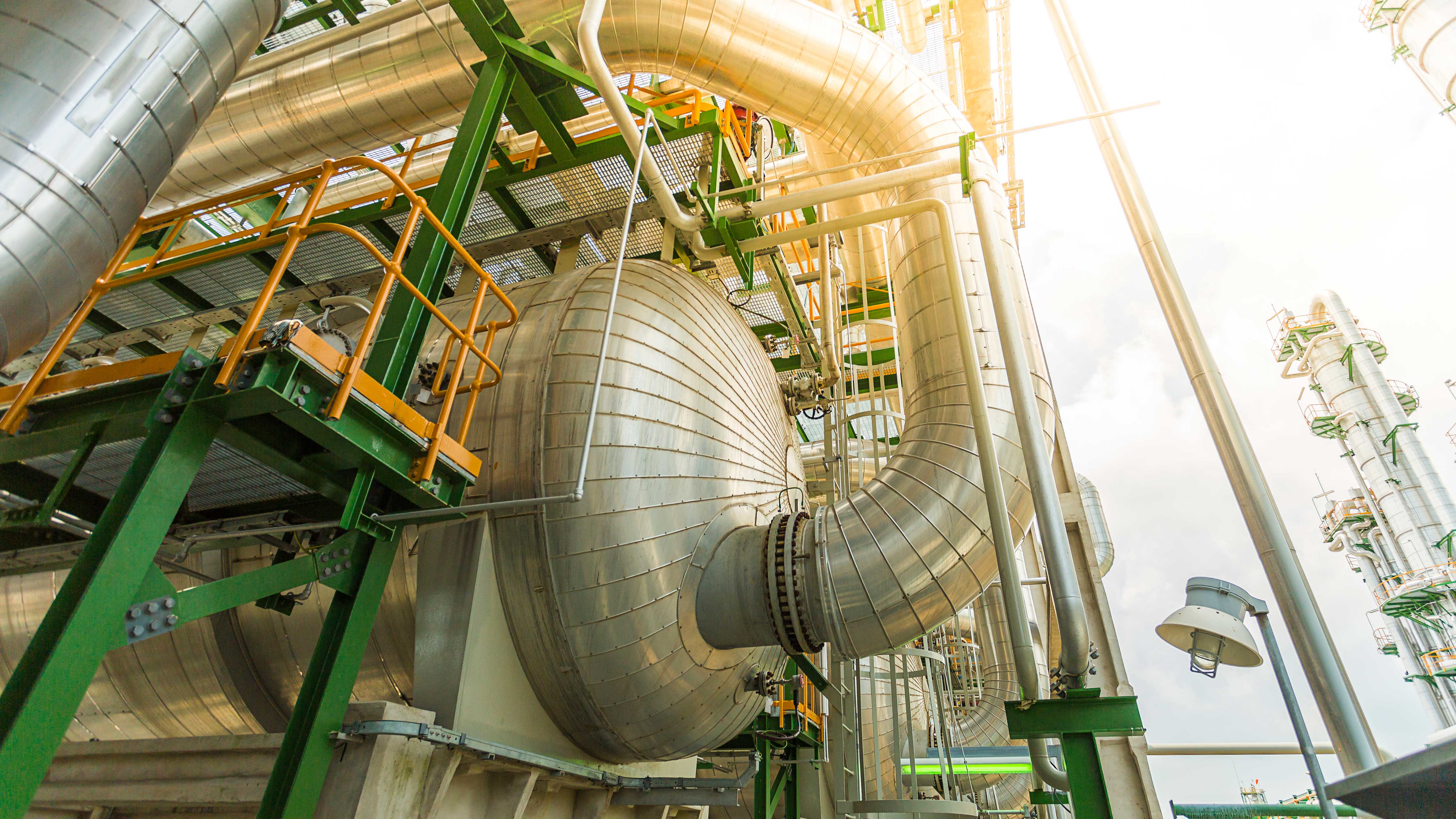 Industrial facility with large metallic pipes, a round tank, and a green support structure under a cloudy sky.