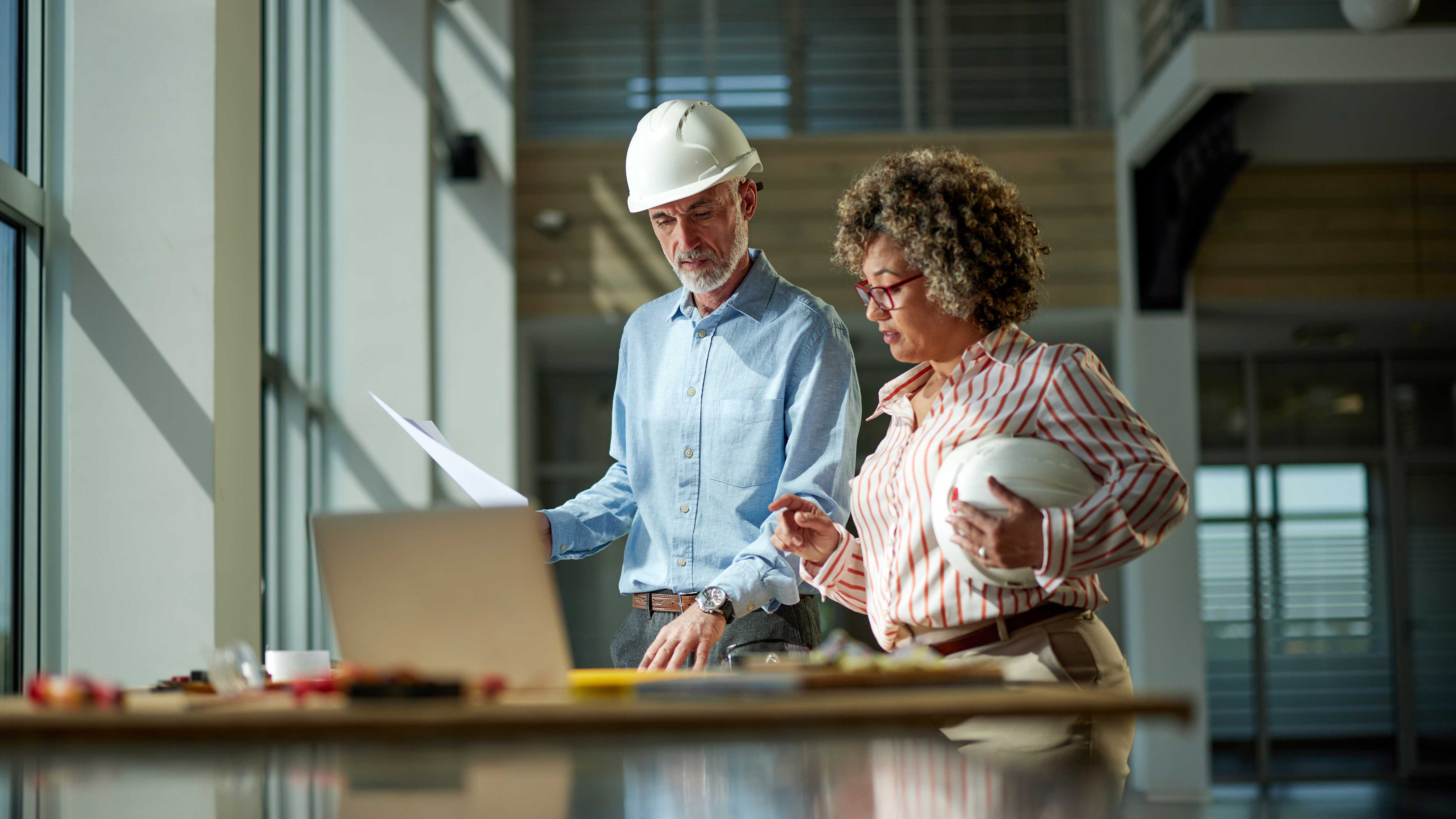 Two professionals discuss plans at a table with a laptop and construction materials in a well-lit office space.