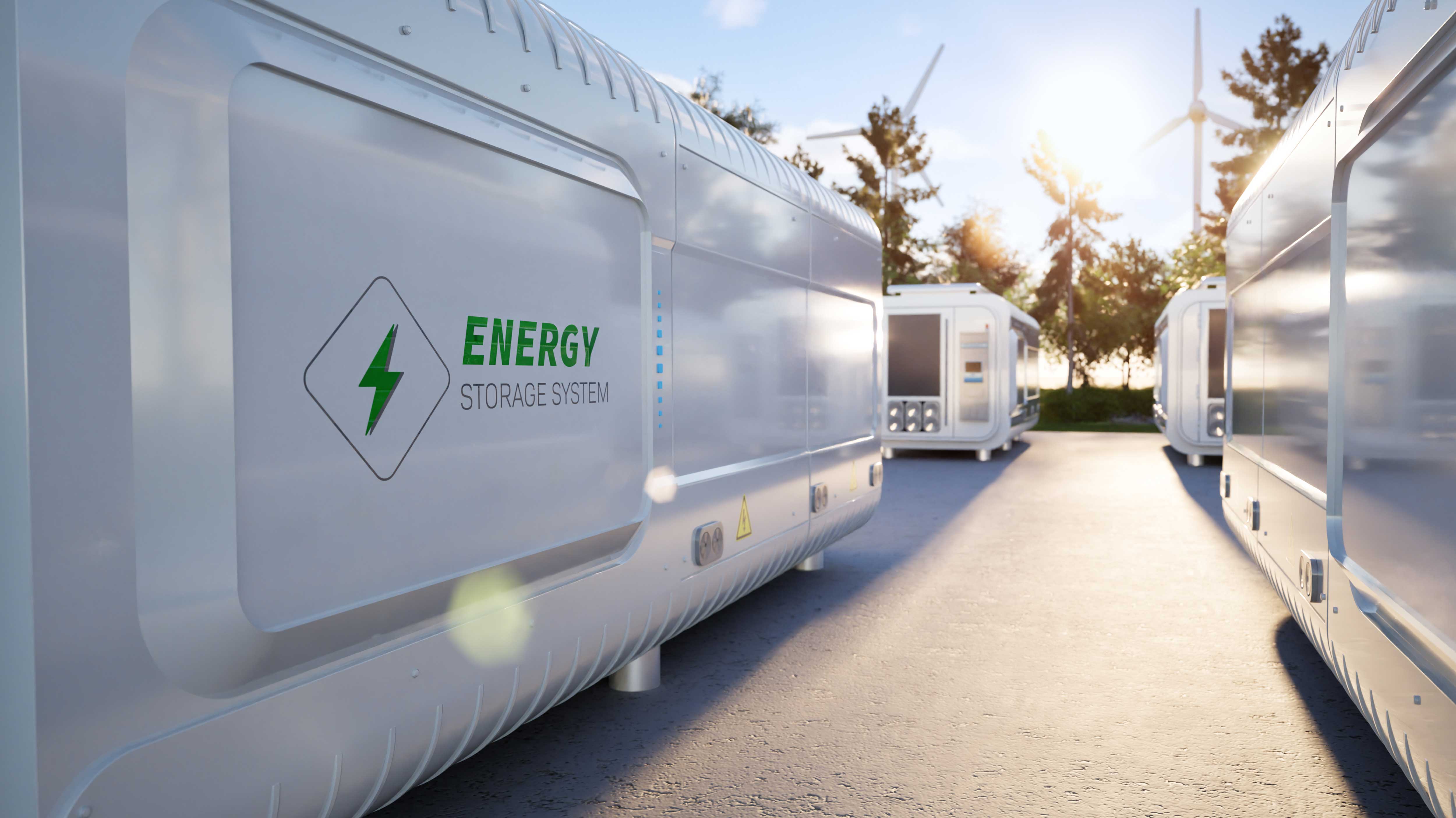 Two energy storage containers are lined up on a paved area, with wind turbines and trees in the background.
