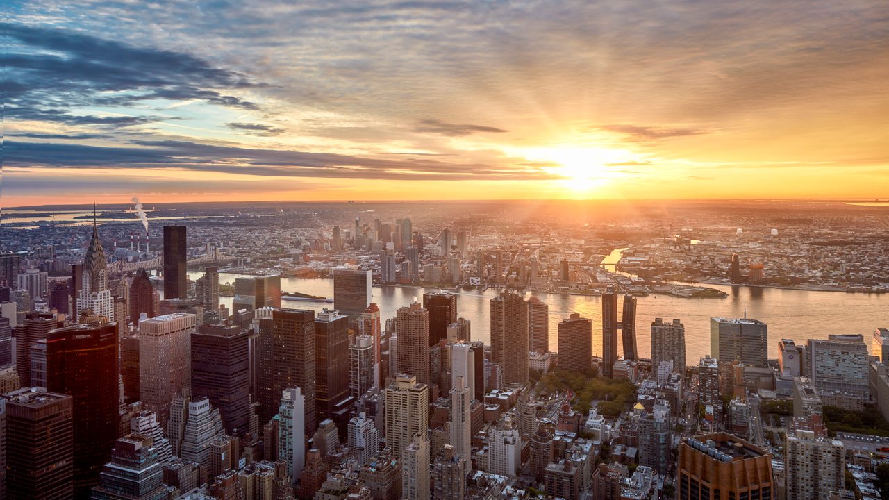 Aerial view of a city skyline at sunset, with buildings and a river reflecting the light.