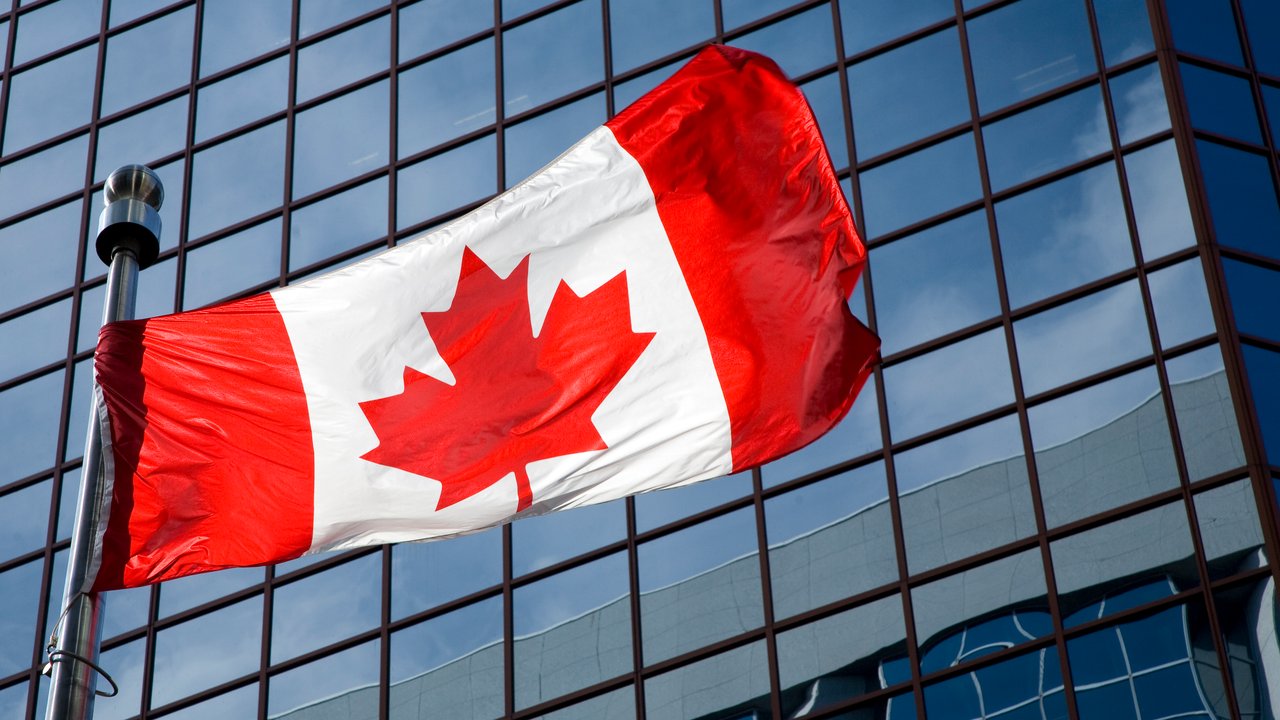A Canadian flag waving in front of a modern glass building reflecting the sky.