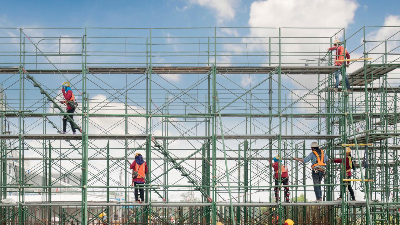 Construction workers on scaffolding, wearing helmets and safety vests, against a blue sky with clouds.