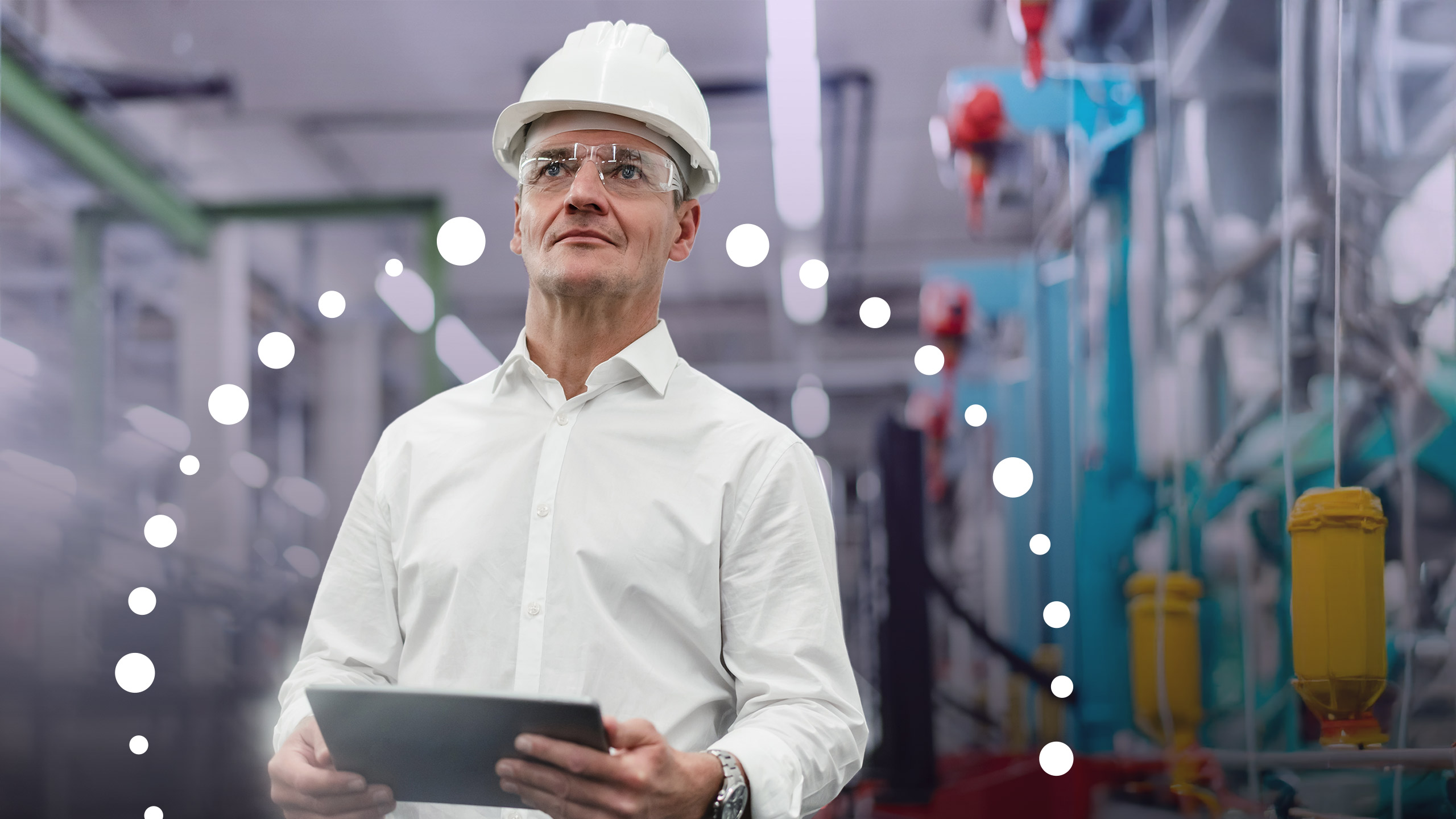 A person in a white shirt and hard hat holds a tablet in a factory setting with machinery in the background.