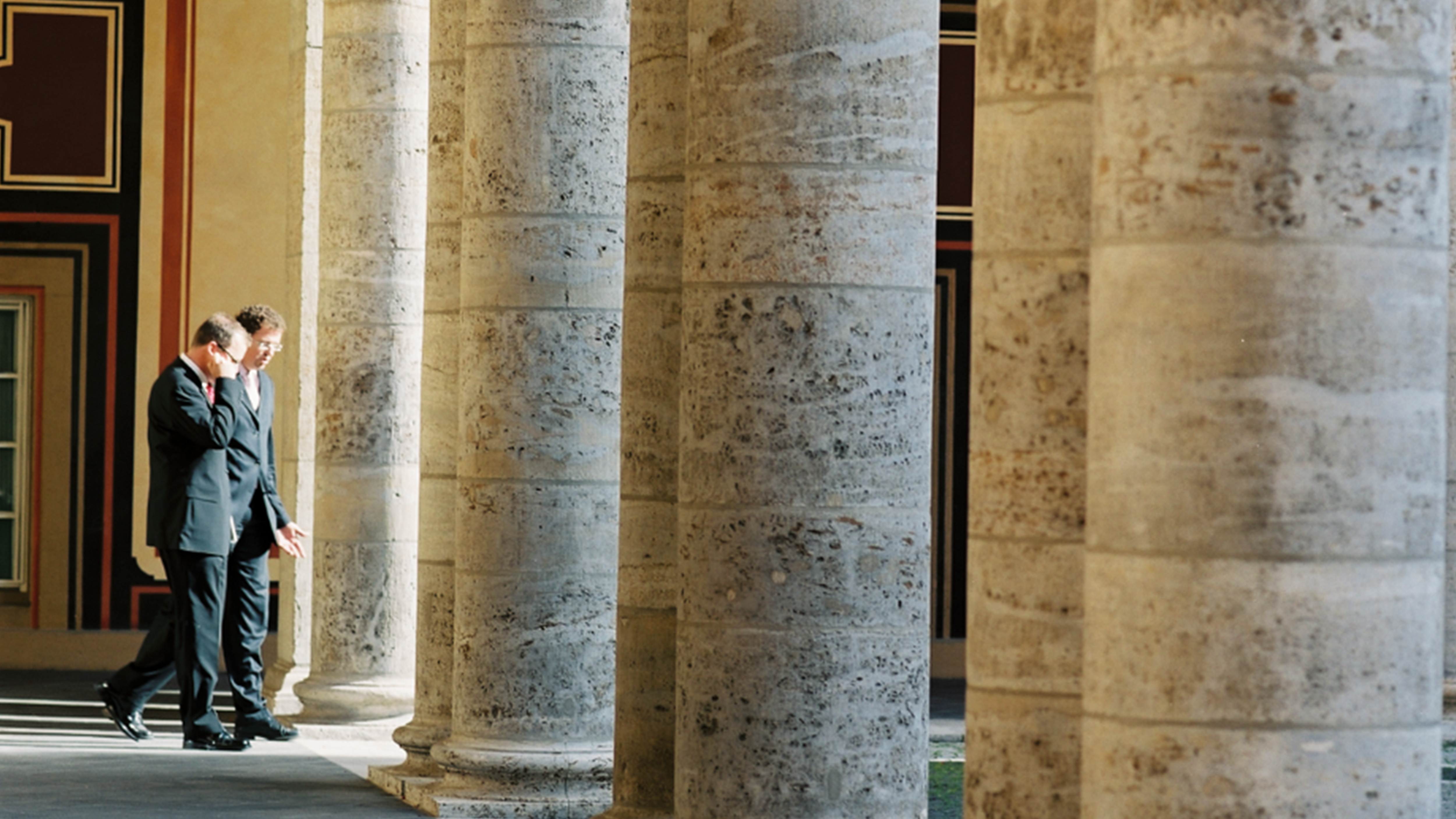 Two men in suits walking by stone columns Two men in suits walk between stone columns in a well-lit corridor.