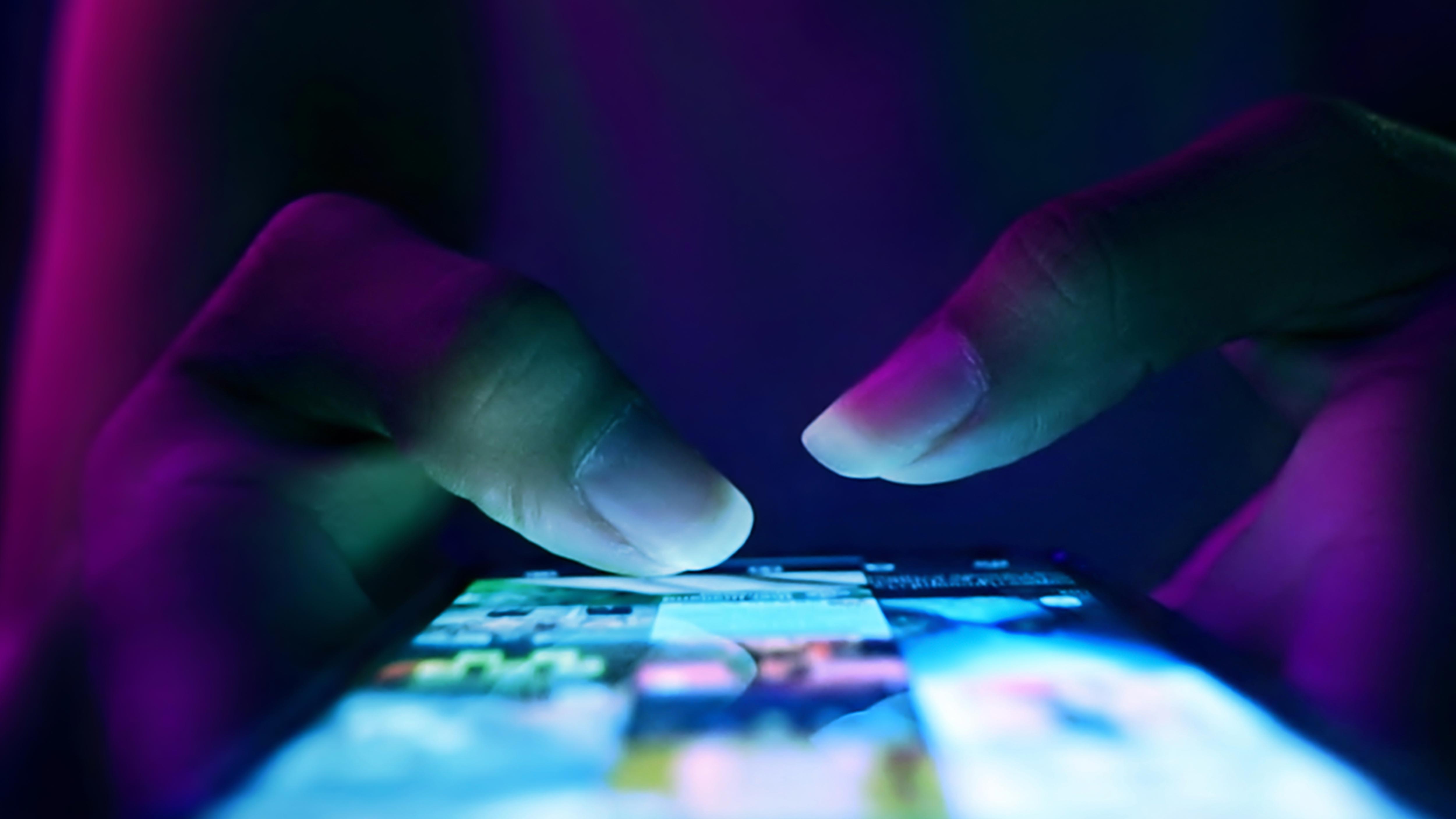 A close-up of hands using a smartphone with a brightly lit screen in a dark environment.