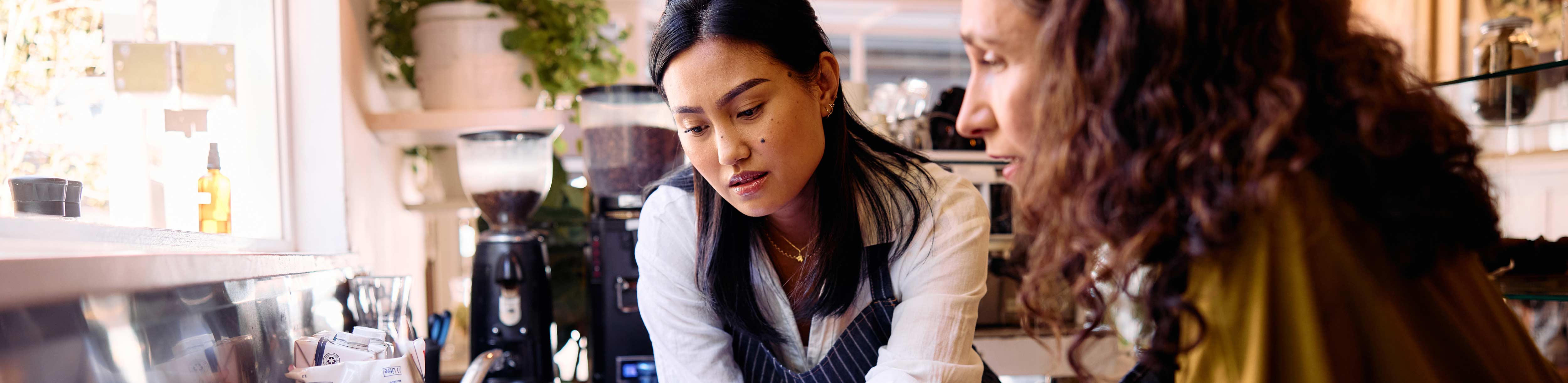 Two women interact at a café counter, with coffee equipment and condiments visible in the background.