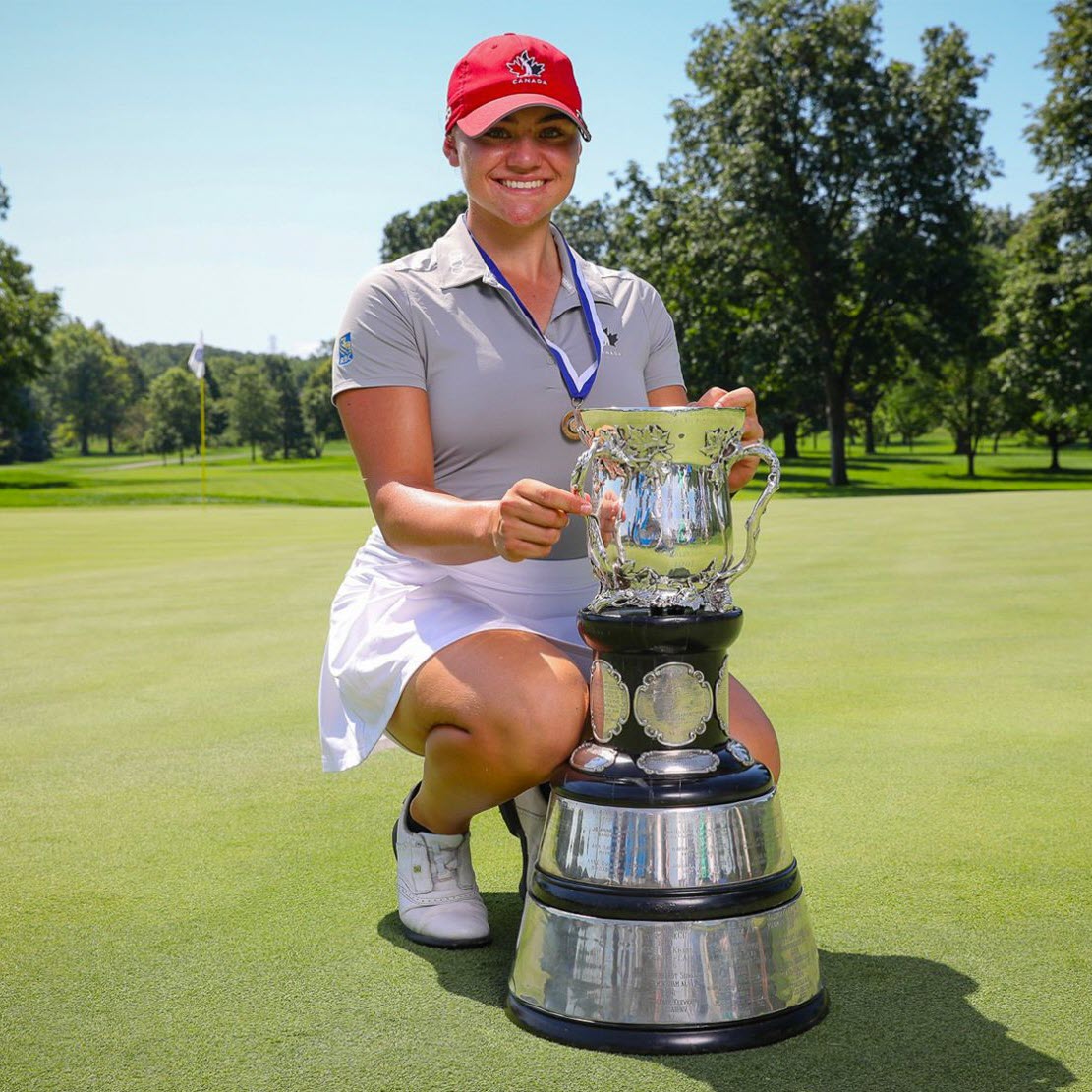 A person kneels on a golf course holding a trophy, wearing a cap and a medal, with trees in the background.