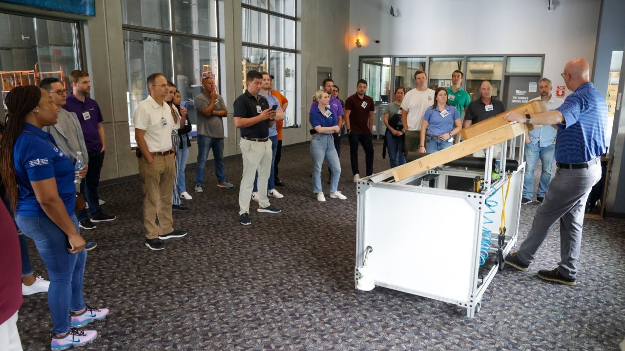 A group of people observes a presentation involving a sloped wooden ramp and a demonstration setup.