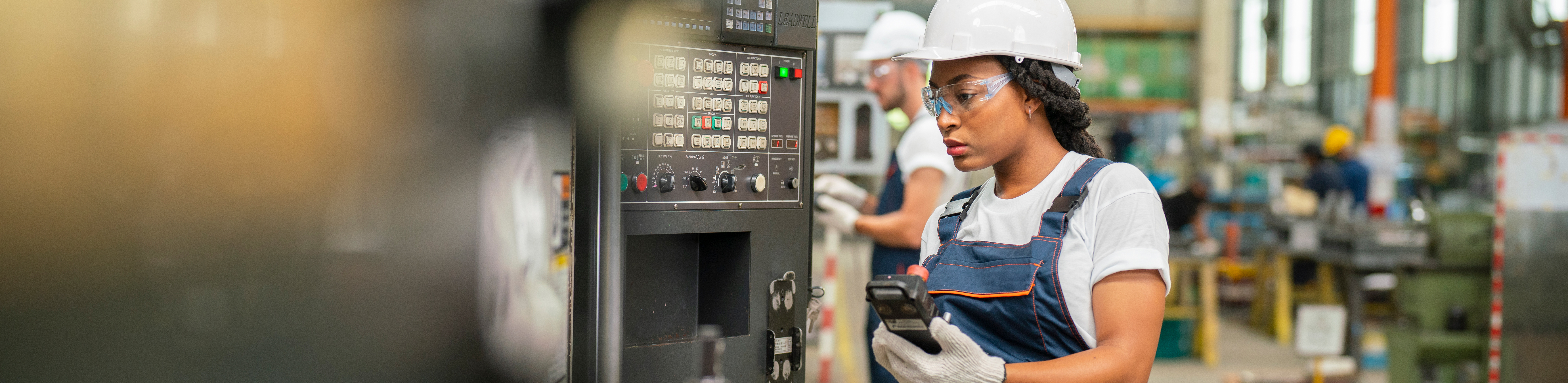 A worker in a factory setting operates machinery, wearing a hard hat and gloves, with equipment in the background.