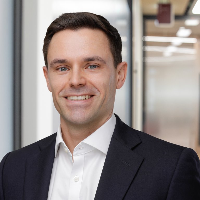 A smiling man in a suit stands in an office setting with blurred backgrounds of glass walls and warm colors.