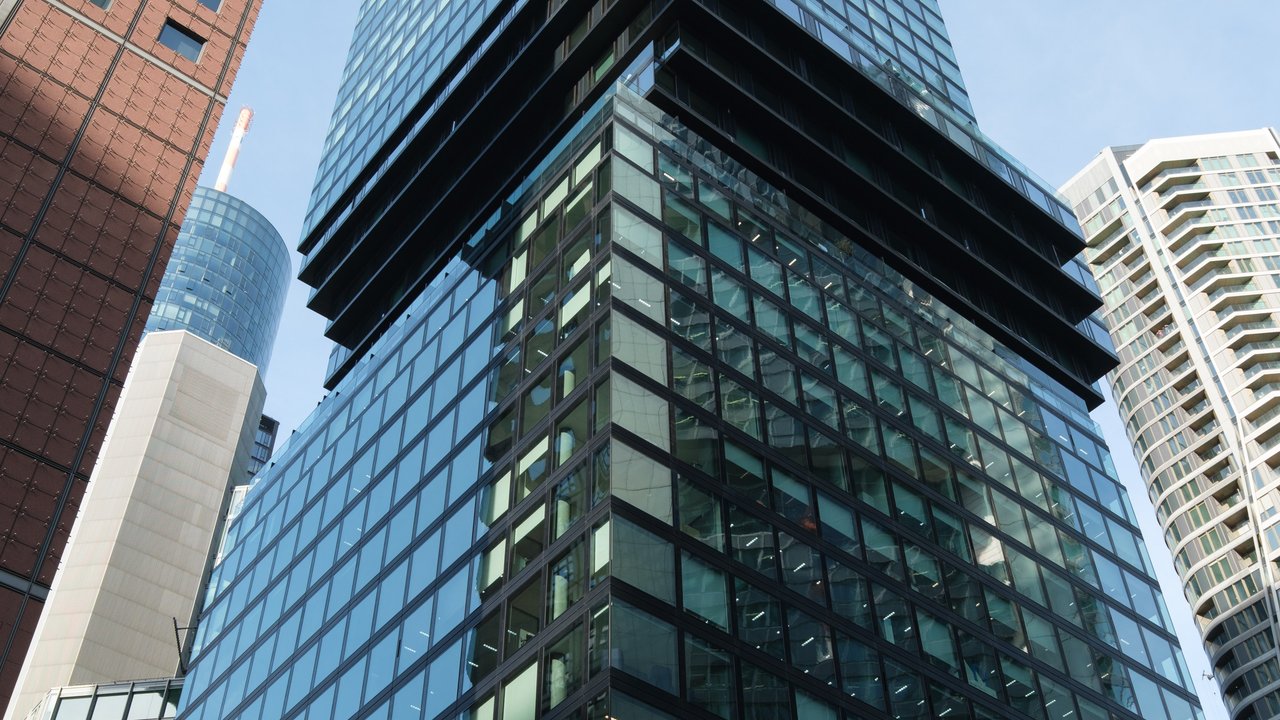 A modern glass skyscraper surrounded by other buildings, viewed from below against a clear sky.