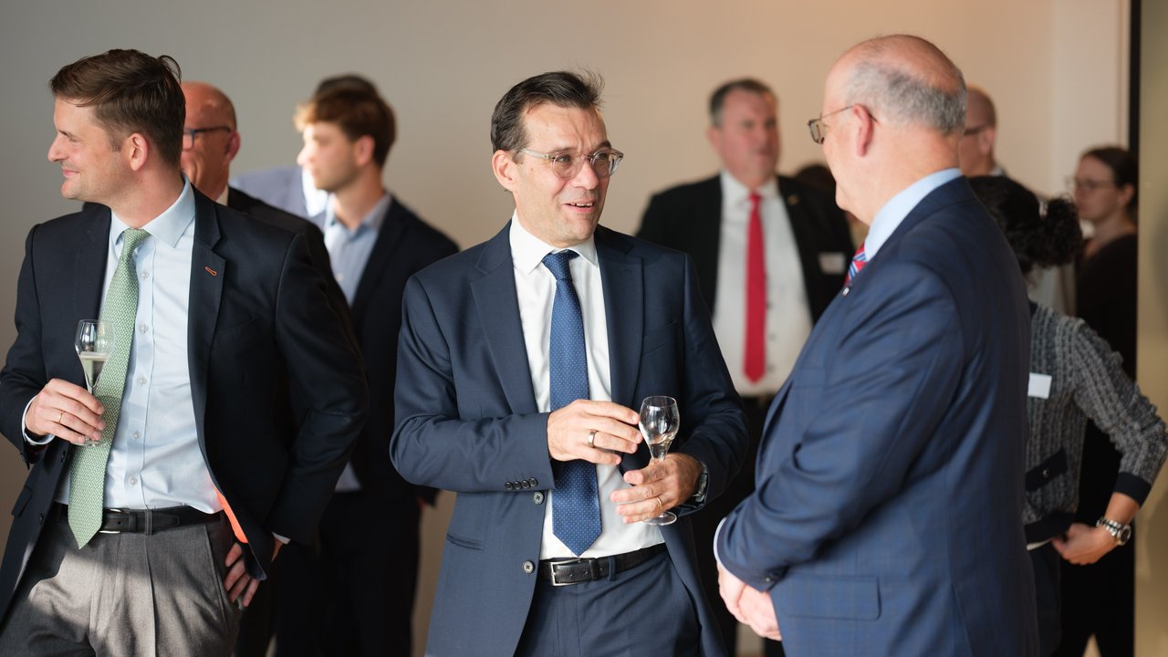 A group of men in business attire conversing at an event, some holding glasses, with a neutral background.