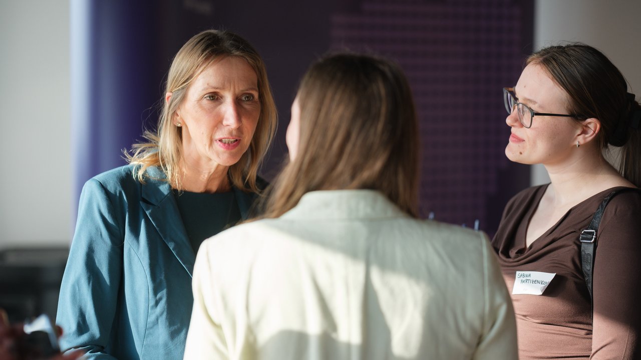 Three women engaged in conversation, one wearing a teal blazer, another in a light jacket, and the third in a brown top.