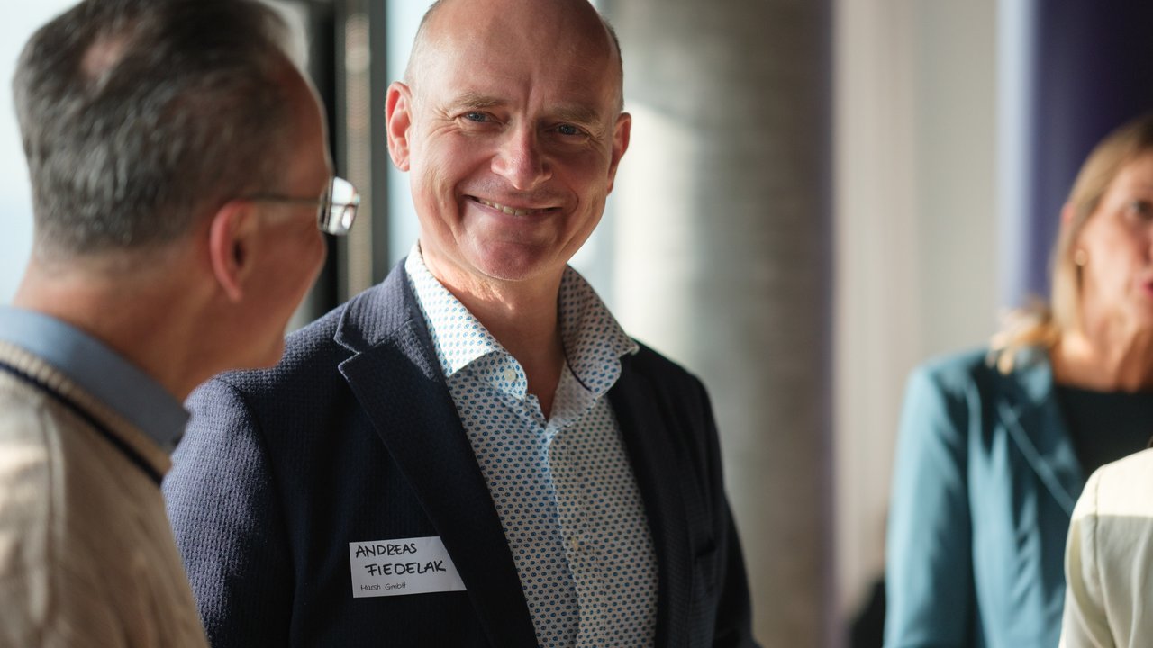 A man in a suit smiles while talking to another man in glasses, with a woman in the background.