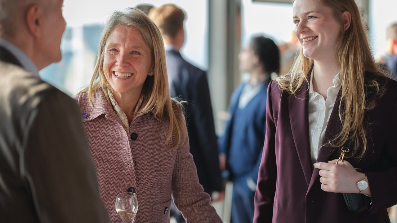 Two women are smiling and talking, one holding a glass, with blurred figures in the background.