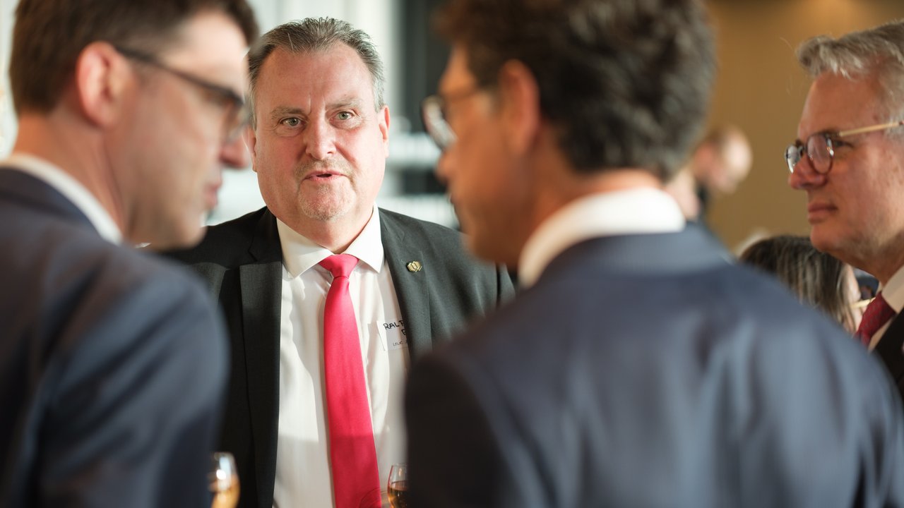 A group of four men in suits conversing at an event, one holding a drink, with a blurred background.