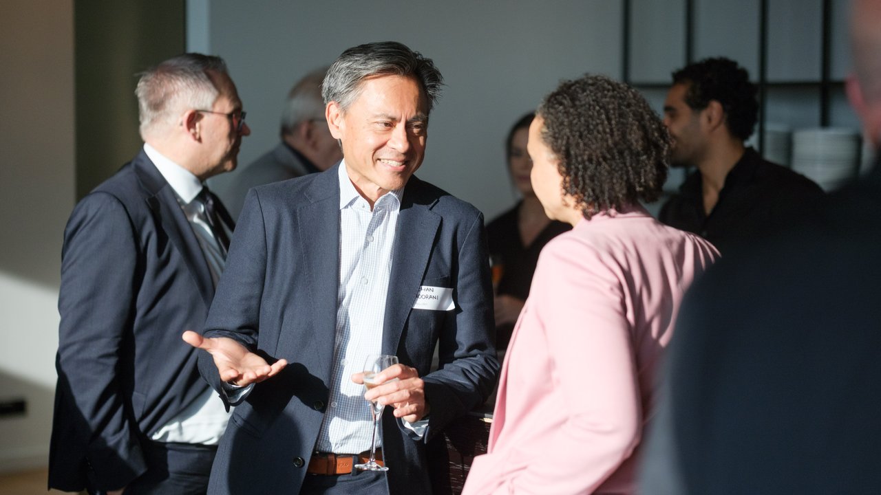 A man in a suit talks with a woman in a pink blazer at a social gathering, with others in the background.