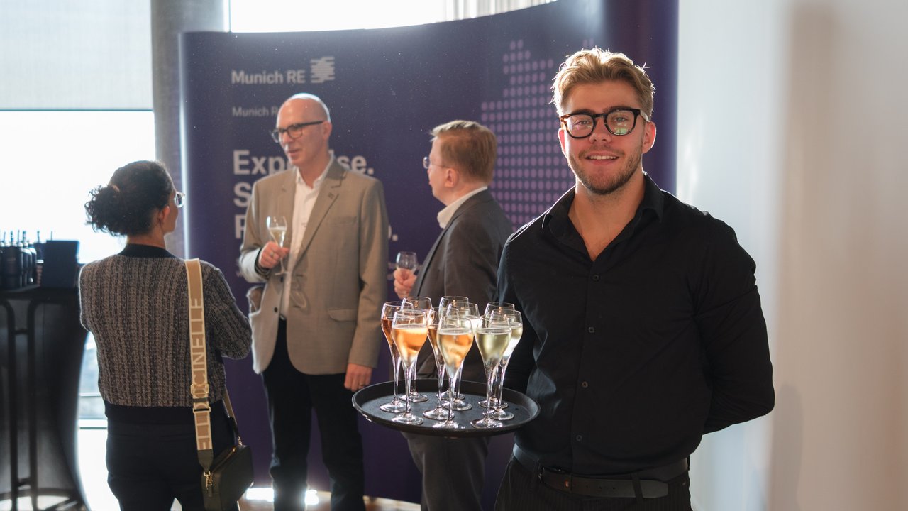 A man in a black shirt holds a tray of drinks, while guests converse in the background near a Munich RE banner.
