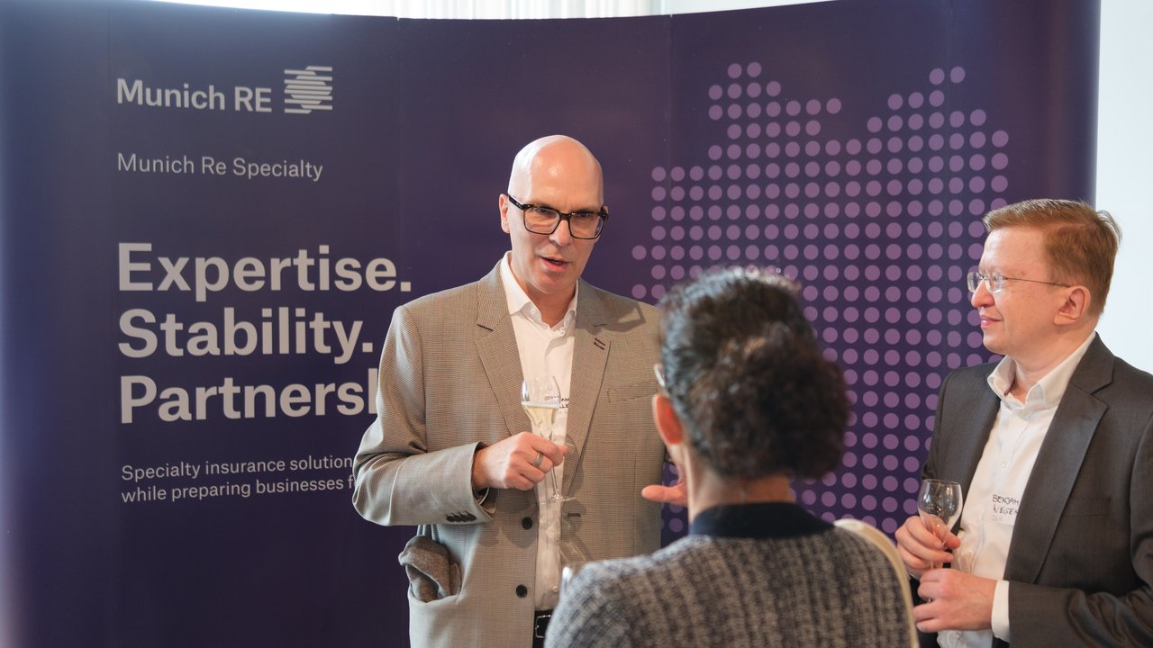 Three people engage in conversation at a Munich Re Specialty event, with a branded backdrop displaying key phrases.