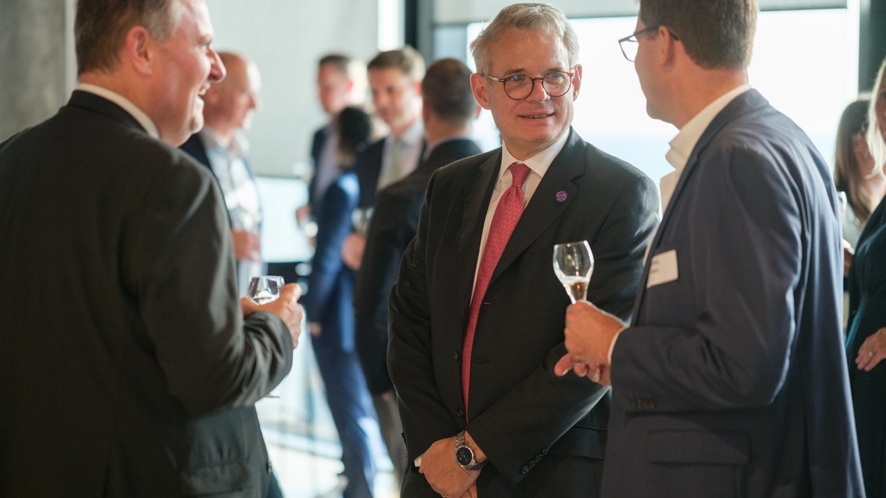A group of people in business attire conversing at an event, holding glasses of drinks.