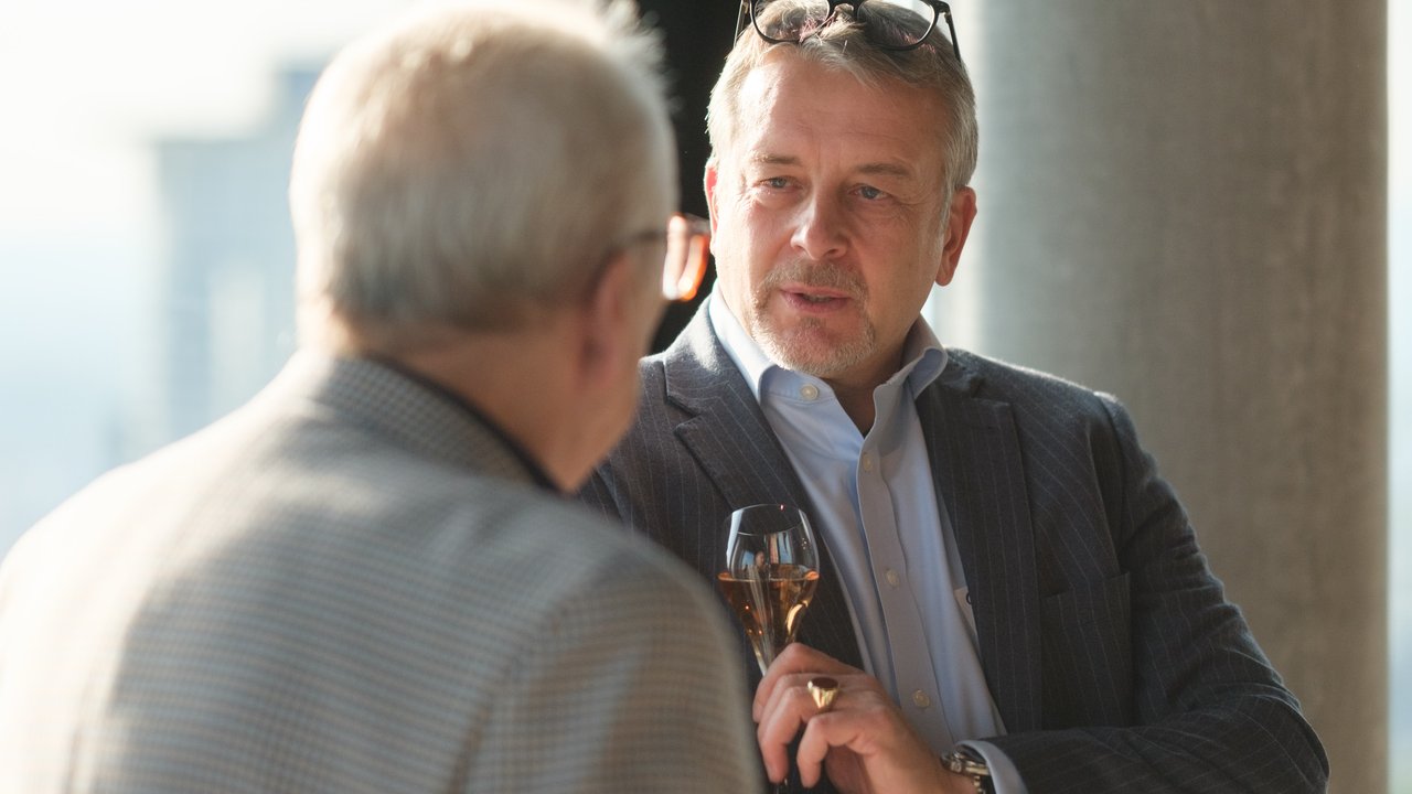 Two men in conversation, one holding a glass of champagne, with a city view in the background.