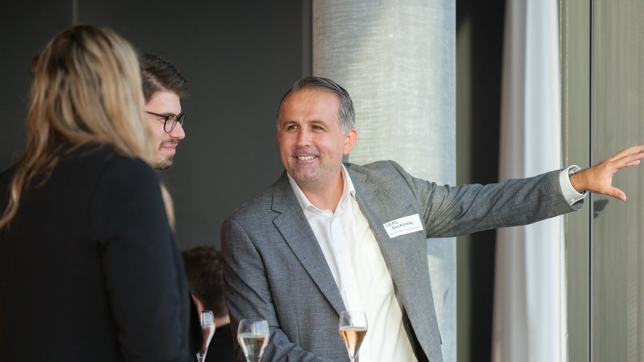 A man in a gray blazer gestures while speaking to two people at a social gathering, with drinks on the table.