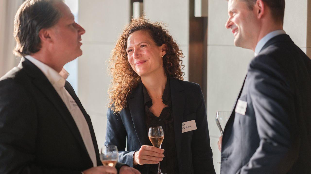 Three people in business attire engage in conversation, holding glasses of champagne in a well-lit indoor setting.