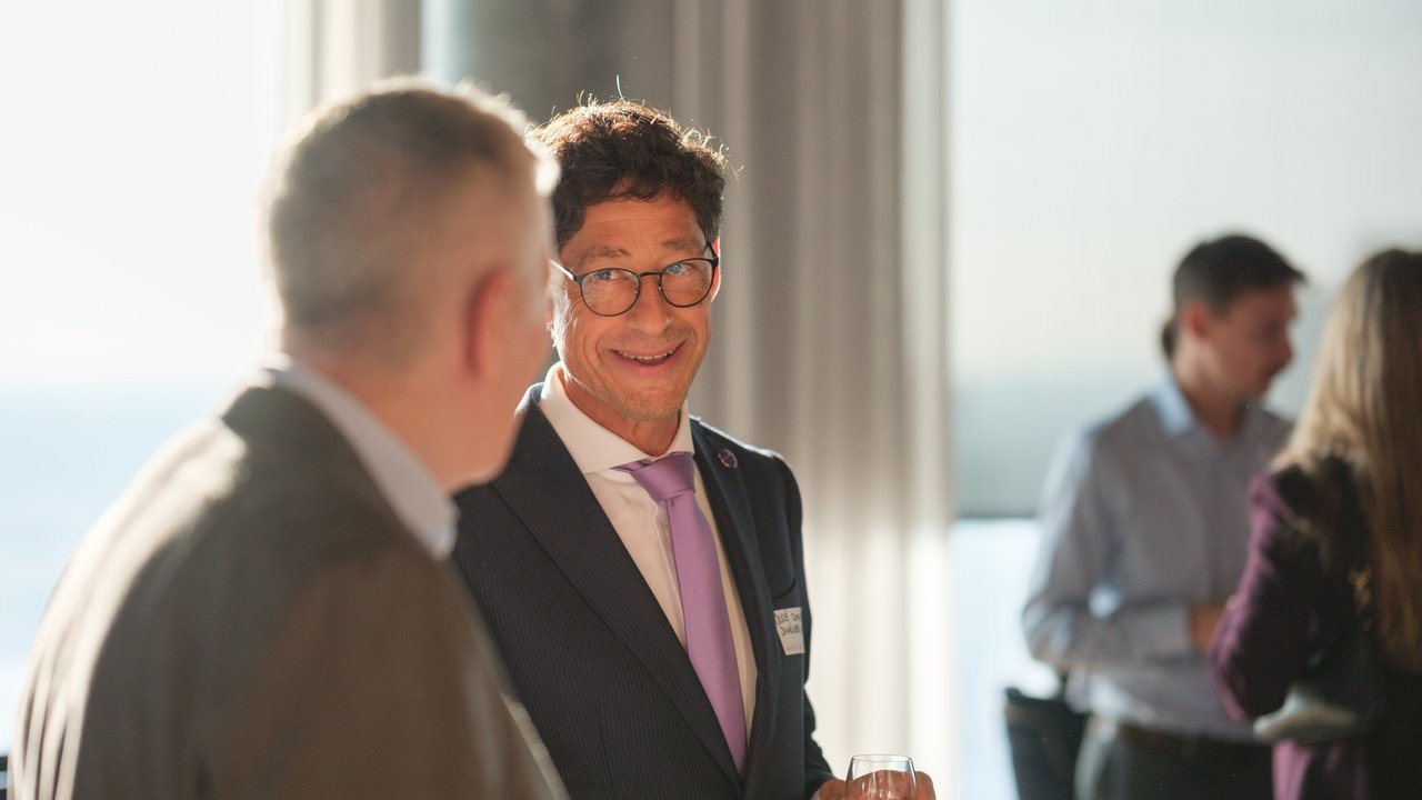 A man in a suit smiles while holding a drink, conversing with another man in a social setting.