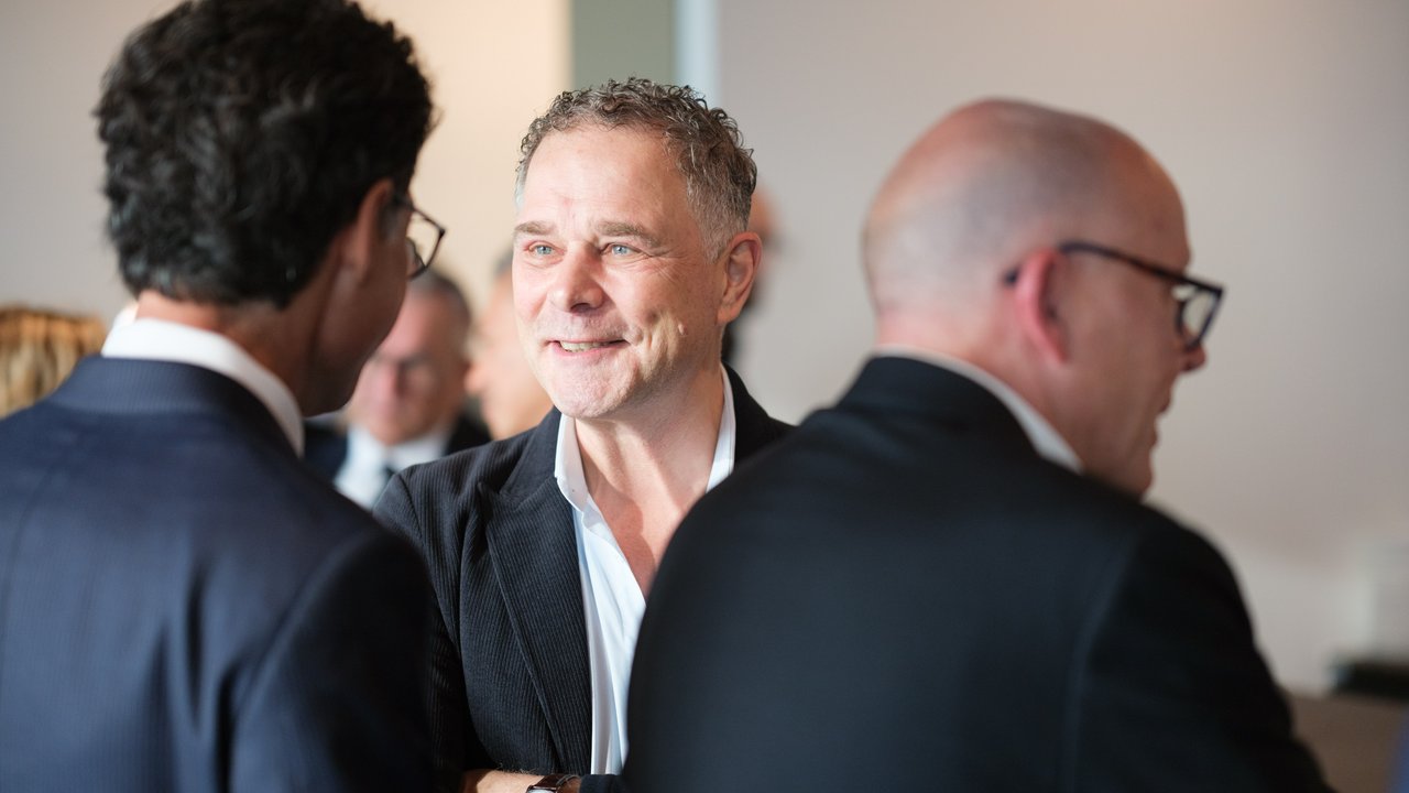 Three men in business attire engage in conversation at a social event, with one smiling and making eye contact.