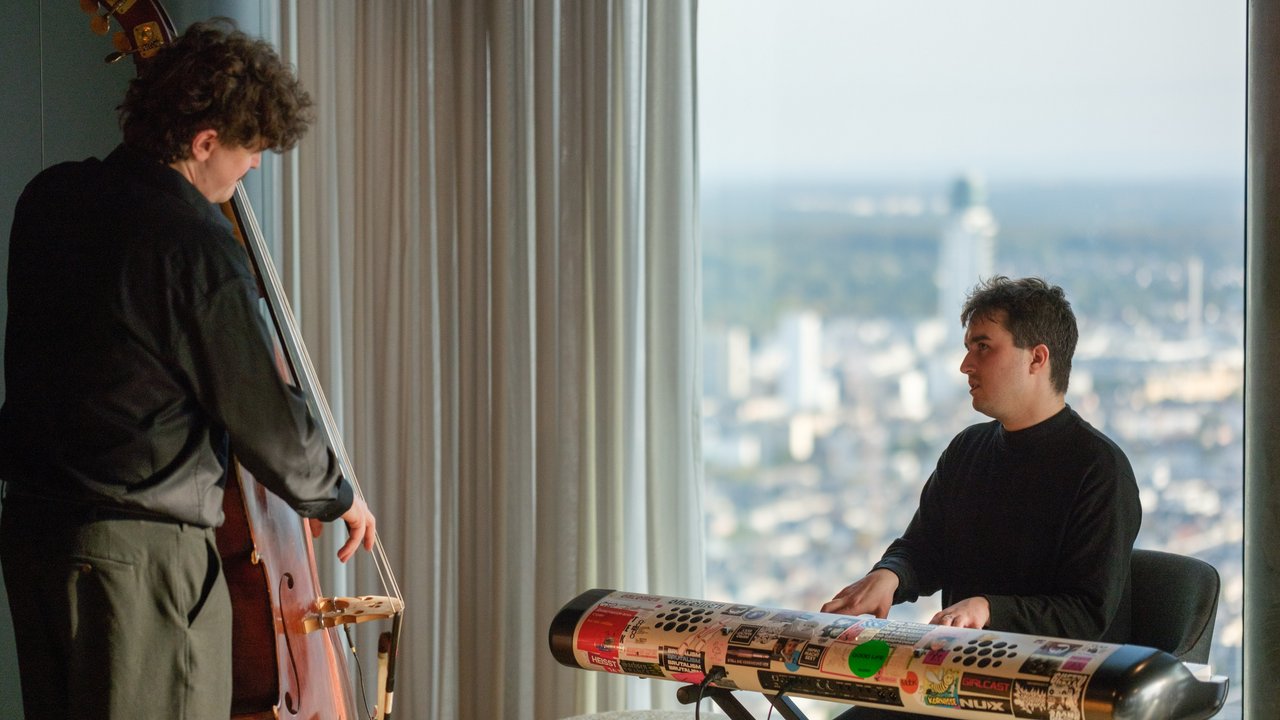 Two musicians perform indoors: one plays a double bass, the other plays a keyboard with stickers, city view behind them.
